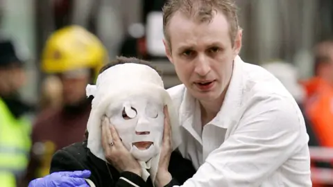 PA Media A woman with a white burn treatment mask over her face. Paul is clutching her with both of his arms around her. He is wearing a white shirt and has brown hair. He looks distressed
