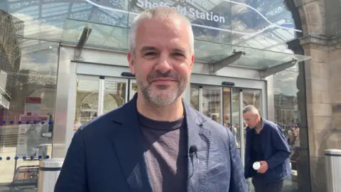 BBC/Naj Modak A man with short grey hair and stubble is wearing a navy blue jacket and dark t-shirt. He is standing outside a railway station with large glass windows in the background