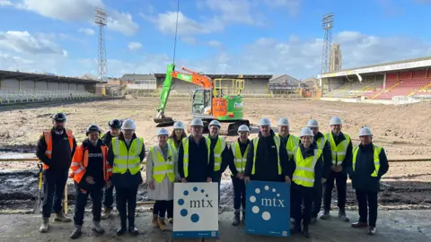 A group of 15 people in hard hats and high vis vests stand on a concrete area. Behind them the pitch has been competely dug up and a digger stands in the middle. The old stands and flood lights can be seen in the background.