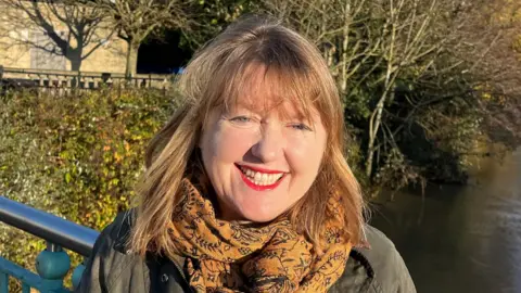 Sarah Gibson smiles at the camera. She wears a gold scarf and dark coloured jacket