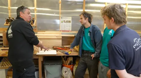 Martin Giles/BBC Three apprentices stand in a workshop. They are watching and listening to Alan Fuller, who is gesturing to something on a workbench. He is wearing a black fleece, with "Pioneer Sailing Trust" on the back. 