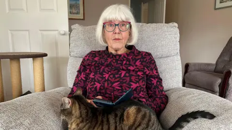 Liz Walton is pictured sitting in an armchair wearing a pink and red patterned blouse with a tabby cat sitting on her lap. She has short white hair with a fringe. Walton is wearing glasses with blue and pink frames.
