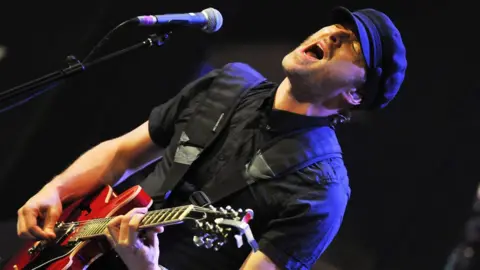 Jez Williams, with stubble, wears a black peaked cap and a black short sleeved shirt as he plays a red guitar and sings into a microphone