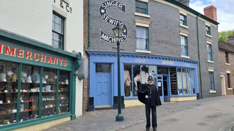 A man is dressed as a Victorian Policeman with a black hat and clock-like coat. He is smiling and waving. He is standing in a Victorian Street with a lamppost next to him. To the left is a green shop front with red sign and kitchen appliances in the window. Behind that is a blue shop front with clothes in the window