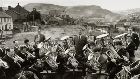 Estate of Evelyn Hofer, courtesy Galerie m Bochum, Germany Mid-Rhondda Workman's Band in Penygraig in 1964: a black and white photo showing a large group of men in suits playing brass instruments, with the valley, hills and housing in the background
