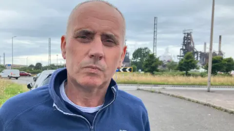 Paul Wagstaffe, wearing a blue jumper standing on the roadside in front of Tata Steel's blast furnaces in Port Talbot, in the background