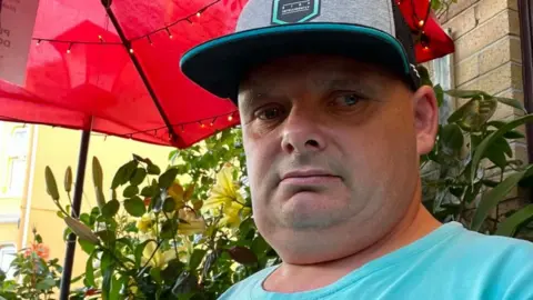 Darren Evans, a man his 50s, wearing a baseball cap and turquoise blue t-shirt. He is sitting under a large red sun umbrella