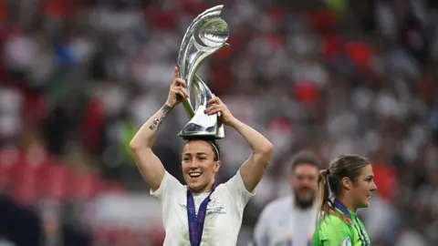Reuters Bronze holding the Women's Euro trophy on her head. It's silver with a glass sphere-like shape at the top. She's smiling and looking off to the side. She's wearing a white England kit and a purple medal.