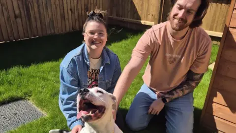 A photo of Charlotte and her partner Ryan kneeling down on grass behind Sasha the white Staffordshire bull terrier, which has its mouth open and its head arched to the sky. Everyone is happy. 