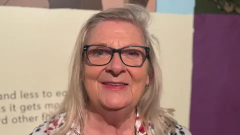 An older lady stands in a museum space. She has long blonde hair and wears dark framed glasses. She is smiling widely and is wearing a brightly coloured floral print shirt. 