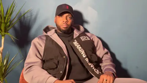 A picture of Rico sitting in a dark red chair in front of a dark blue wall, next to a plant on the left. He wears a black jumper under grey tracksuit with a black gilet jacket on top with the white text ‘Directed by Rico’ on the breast and ‘Making Movies’ down the zip seam.