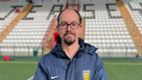 Tim Pryor lleva un abrigo azul marino con el logo de Nike y el escudo de la FA de Jersey en el otro lado. Luce una barba de chivo oscura, gafas y cabello castaño. Detrás de él se ven las gradas de un estadio de fútbol, blancas con la palabra Jersey escrita en negro.
