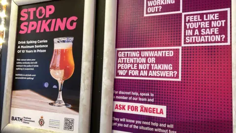 A series of posters on a bar wall showing a warning for spiking and to Ask for Angela.