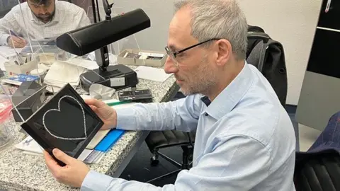 Peter Jackson Peter Jackson seated at a desk in a pale blue shirt looking down at a black box with diamonds in a heart shape that he is holding in his hands