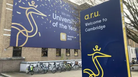 Brian Farmer/BBC Sign for Anglia Ruskin University. It is blue with a yellow heron insignia on it. Outside the university building are rows of bicycles.