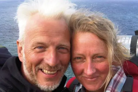 A woman, wearing a red jacket, poses for a selfie with her husband who has grey hair and is wearing a black jacket. In the distance is the sea. 