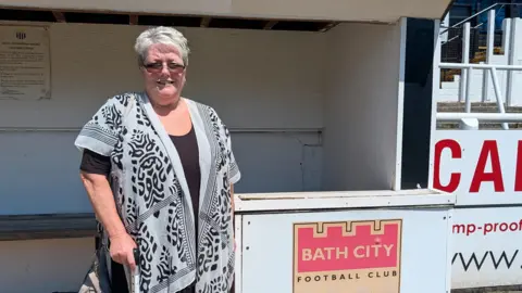 Bath City FC fan Jayne Smith standing by the stands at Bath City FC. There is a logo saying Bath City Football Club on a board to her left. She is wearing a black and white, long top and has short hair and glasses.