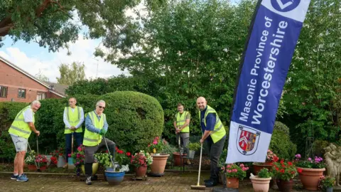 A group of five men in yellow high viz jackets carry out work in a garden area. The men pose next to a purple banner that reads: "Masonic Province of Worcestershire." 
