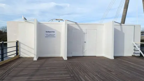 Kaleigh Watterson/BBC A seagull sits on the white boards across the wooden Southport Pier, with a notice stating it has been shut due to safety issues