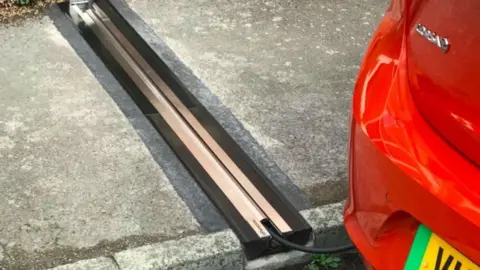 A gully cutting across a cable with a metal cover and a wire protruding and connected to the back of a red electric car.