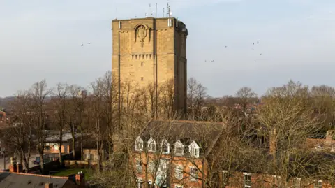 An aerial shot of the tower in winter - the trees are bare and there are birds flying above the buildings below it. It is built of brick and stands nearly three times the height of the three story building below it.