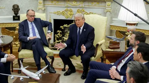 Friedrich Merz, Germany's chancellor, left, and US President Donald Trump during a meeting in the Oval Office of the White House. Both sit on gold fabric chairs and are flanked by staff and aides