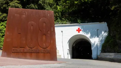 The entrance to Jersey War Tunnels, there is a large metal sign saying 'german underground hospital' and a white tunnel behind it.