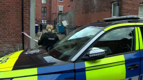 A blonde police officer stands near a police car