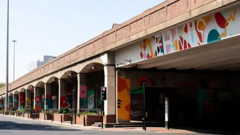 Rachel Deakin A large road has had the underpass decorated with brightly coloured abstract shapes. The word "Erimus" can be read in white along the underside of a bridge. Every pillar holding up the road has been painted with a variety of shapes and colours from pink spots to checkerboard patterns.