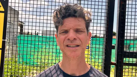 James Allchurch is seen in a dark t-shirt stands in front of a fenced construction site, with warning signs and green netting visible behind him.