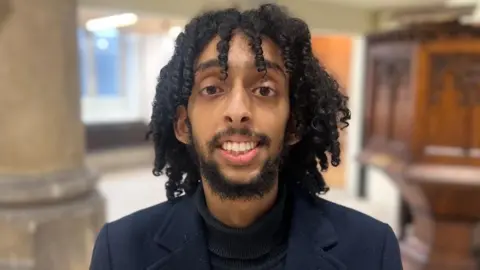 Haitam Amudi is smiling at the camera. He has black shoulder-length hair with a beard and mustache. He's wearing a black jumper and a navy blazer. 