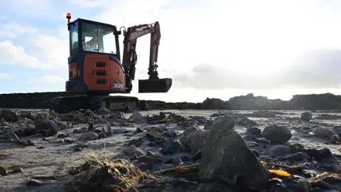Pacemaker A digger sits on a beach which is strewn with debris. There are large rocks, the remains of a collapsed sea wall everywhere
