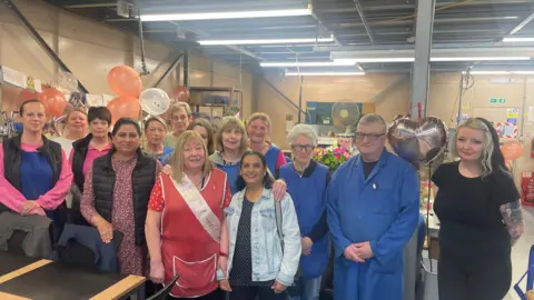 A group photo with Elaine at the front surrounded by her colleagues, some of which are wearing blue overalls. In the background is an industrial warehouse with pink balloons pinned up.