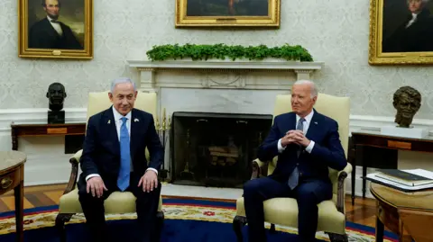 Reuters Benjamin Netanyahu sits with his hands on his knees in the oval office, wearing a blue tight and dark suit. Joe Biden sits to his left, wearing a navy suit with clasped hands. 