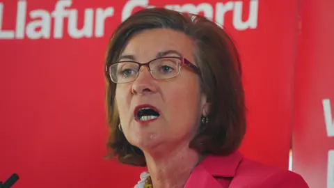 PA Media Eluned Morgan wearing a red jacket in front of a red Welsh Labour banner, with Llafur Cymru written in Welsh behind her