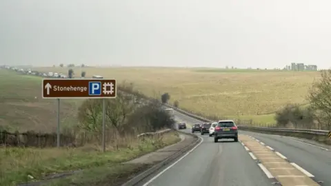 Getty Images A trunk road (A303) going past a Stonehenge parking sign with the stone monument in the background. Trees and grass line either side of the road and there is traffic queuing in the distance under a misty grey sky.