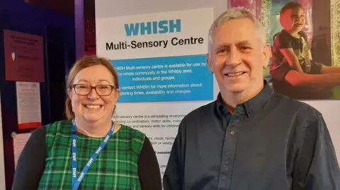 LDRS A woman wearing a green tartan dress and a blue staff lanyard stands smiling beside a man with grey hair wearing a dark shirt. Behind them is a picture of a giggling toddler in a multi-sensory room full of bright lights, beside a sign reading "WHISH multi-sensory centre".