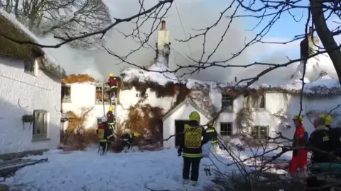 The picture shows a group of firefighters tackling a major blaze at a traditional thatched-roof building. Thick smoke is billowing from the roof, which appears to be severely damaged and partially collapsed. Several firefighters in bright yellow helmets and high-visibility jackets are using ladders and hoses to control the fire, while foam or water covers the ground in front of the building. The words “Ring of Bells” are visible on the front wall.