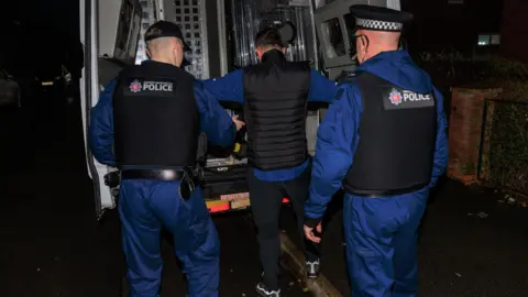 Greater Manchester Police A man wearing a blue long sleeved top under a black bodywarmer and black jogging bottoms is put in the back of a police van by two officers