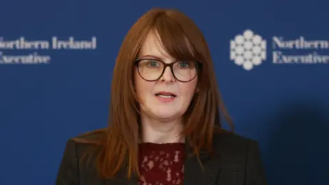 Close photo of Caoimhe Archibald, red haired woman with dark glasses against a blue NI Executive backdrop, wearing a red lacy top and a black suit blazer