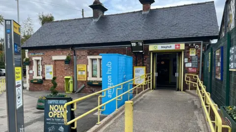 Ian Haslam / BBC The entrance to Maghull railway station, with a sign for The Coffee Carriage coffee shop outside and hanging baskets with flowers on the wall. A planter with flowers can also be seen to the side of the ramped entrance.