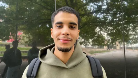 Yassim in a green hoodie standing in front of a black reflective wall