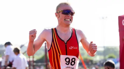 Vannin Photos Corrin Leeming grits his teeth as he crosses a finish line. He holds both hands up by his sides in fists. He wears a red vest and wraparound sunglasses.