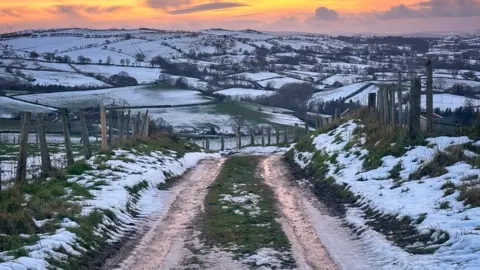 Ruth Davies / Weather Watchers A road with snow either side, the background is the sun setting over snow covered fields. 