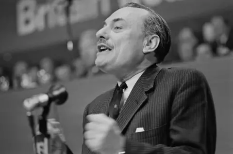 Getty Images A black and white image of a man with a moustache wearing a shirt, tie and suit, speaking into a microphone.