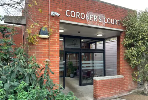 BBC/Harry Low Entrance to East London Coroner's Court shows red brick building, white lettered signs above entrance, a black automatic glass door, and green foliage around the entrance