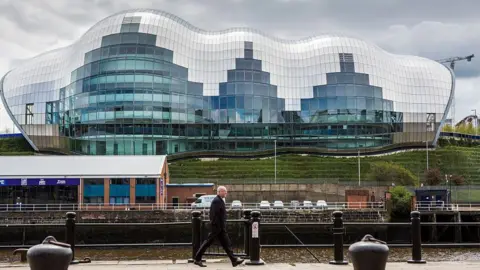 Getty Images The Glasshouse International Centre for Music viewed from Newcastle Quayside. A large building, it has a futuristic shape and is made up of hundreds of glass panels. In the foreground, a man is walking along the Quayside towards to the right of the photo.
