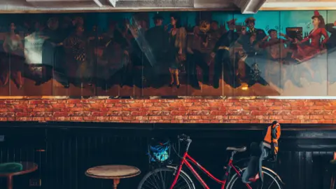 Dom Moore and Nudge Mural inside former club The Clipper. The artwork depicts visitors to the pub. It is displayed on a red brick wall. A red bike has been propped up against the wall under the painting, in front of a black wall.