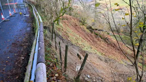 Westmorland and Furness Council Crash barriers at the side of the road where the steep landslip has occurred - the soil is a russet colour surrounded by bare trees. 
