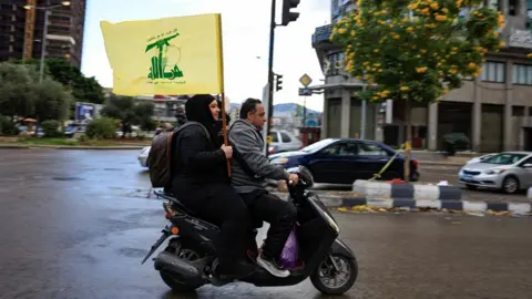 A side view of a woman carries a Hezbollah flag while riding pillion on the back of a motor scooter driven by a man on a street in Beirut